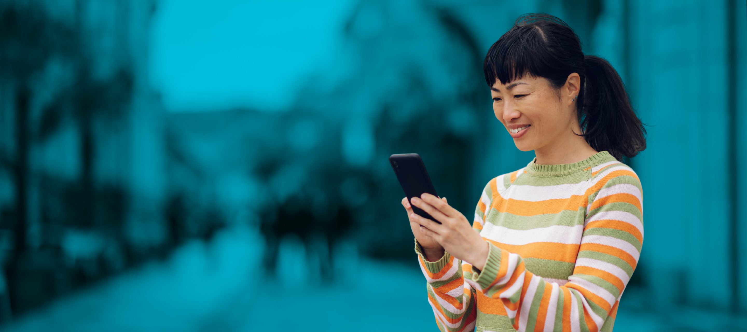 A woman with dark hair, wearing a colourful striped sweater, smiles while using her smartphone to pay for parking.