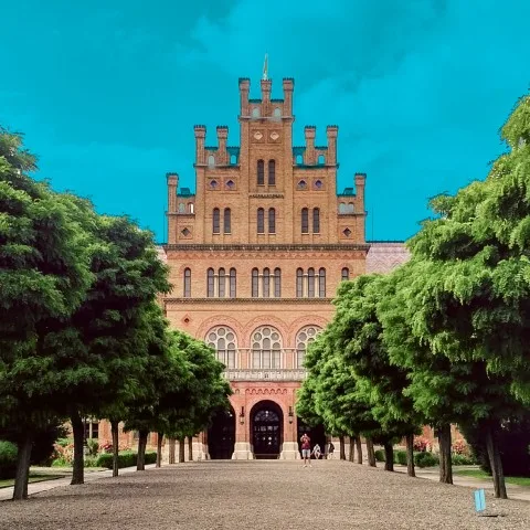 Historic building with a grand architectural style, featuring towers and ornate details, with a pathway lined by neatly trimmed trees leading up to its entrance.