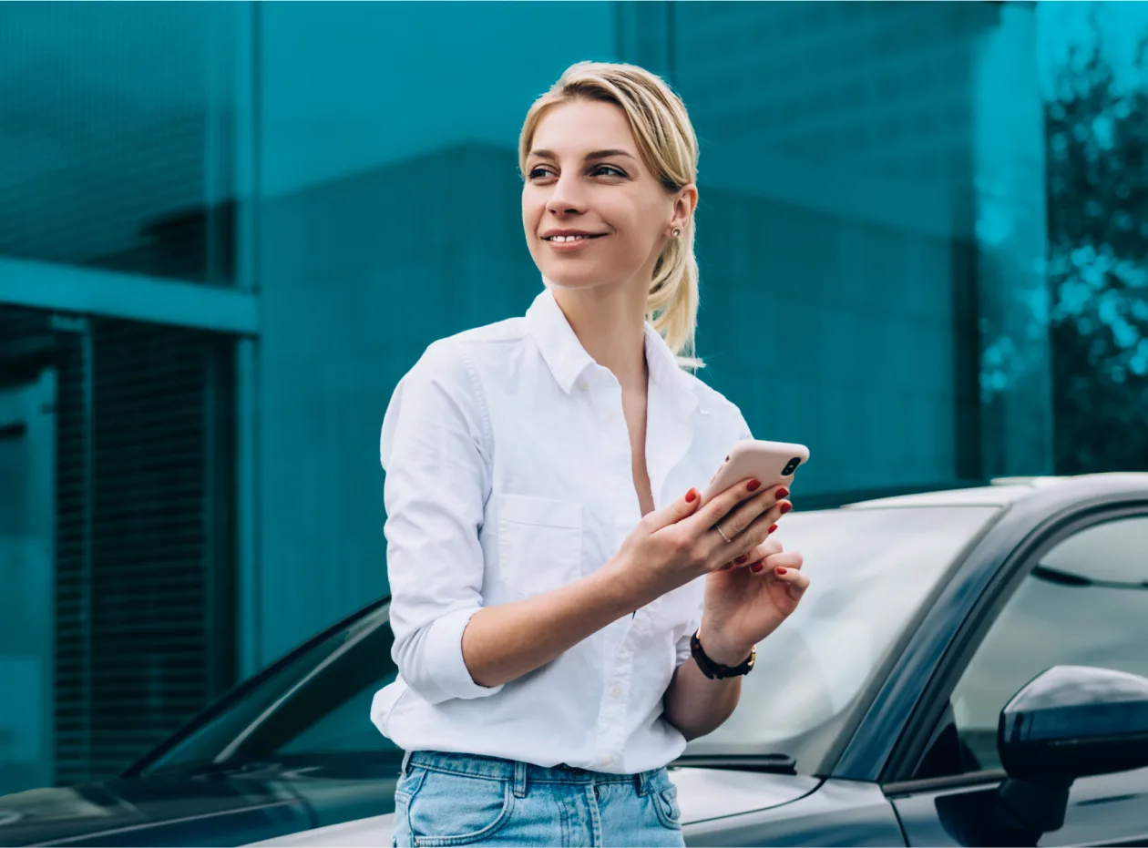 A smiling woman in a white shirt holding a smartphone, standing next to a car in front of a modern glass building.