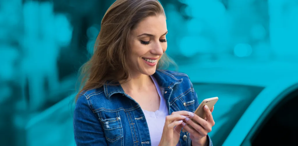 A woman wearing a denim jacket is smiling while using her smartphone in a parking lot.