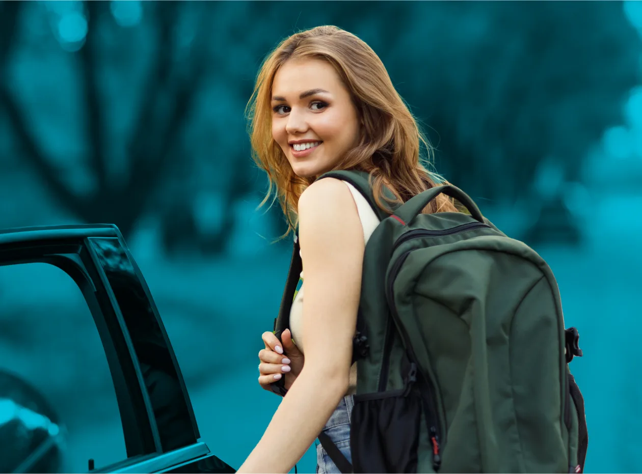 A young woman with blonde hair, wearing a large green backpack, smiles as she opens the door of a car.