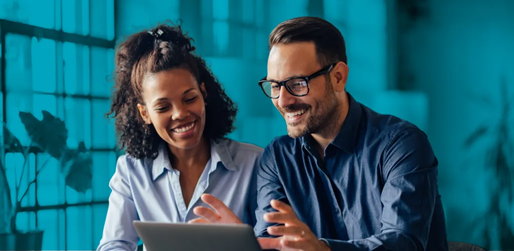 A man and a woman are smiling and looking at a laptop together in a brightly lit office space.