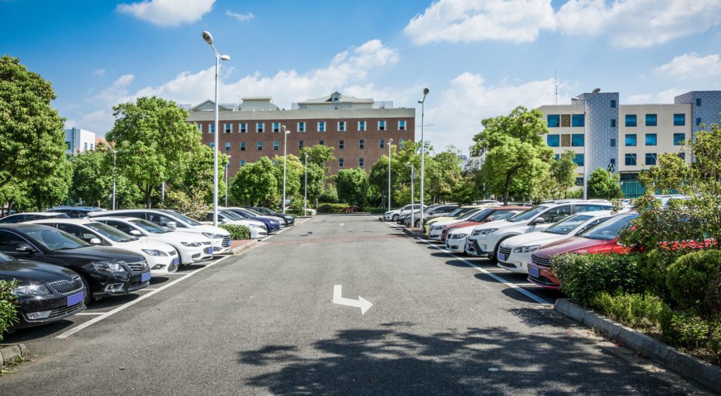 An outdoor parking lot with cars parked on both sides and a white arrow in the central lane, surrounded by trees and buildings in the background.