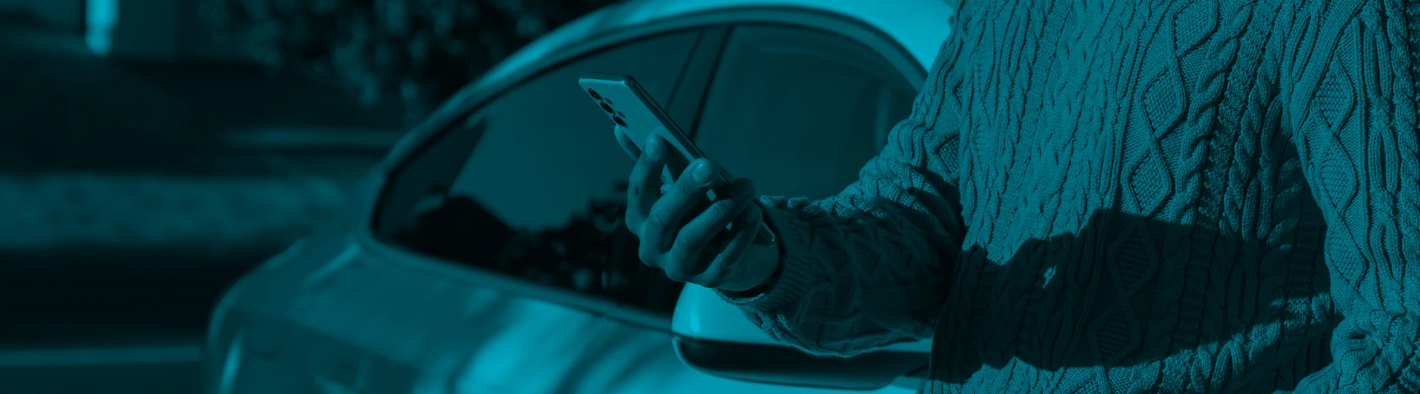 A person wearing a sweater and holding a phone stands near a car.