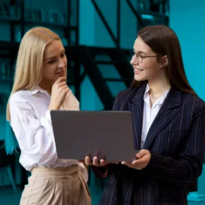 Two women are standing together in a professional setting, one holding a laptop and explaining something while the other listens thoughtfully.