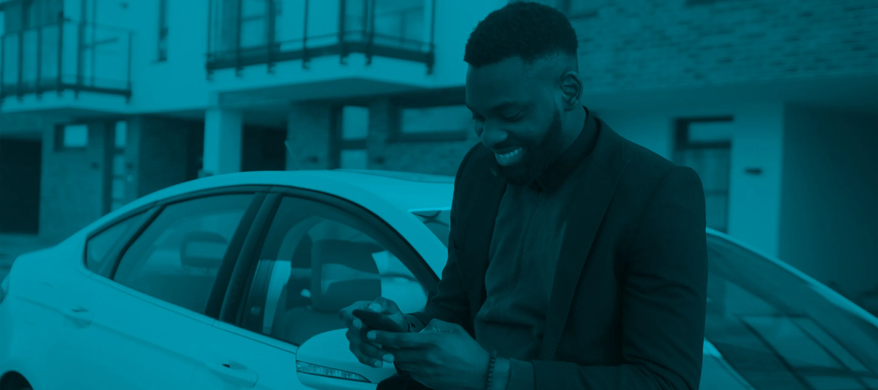 Man smiling while looking at his phone, standing next to a parked car in an urban setting with buildings in the background.