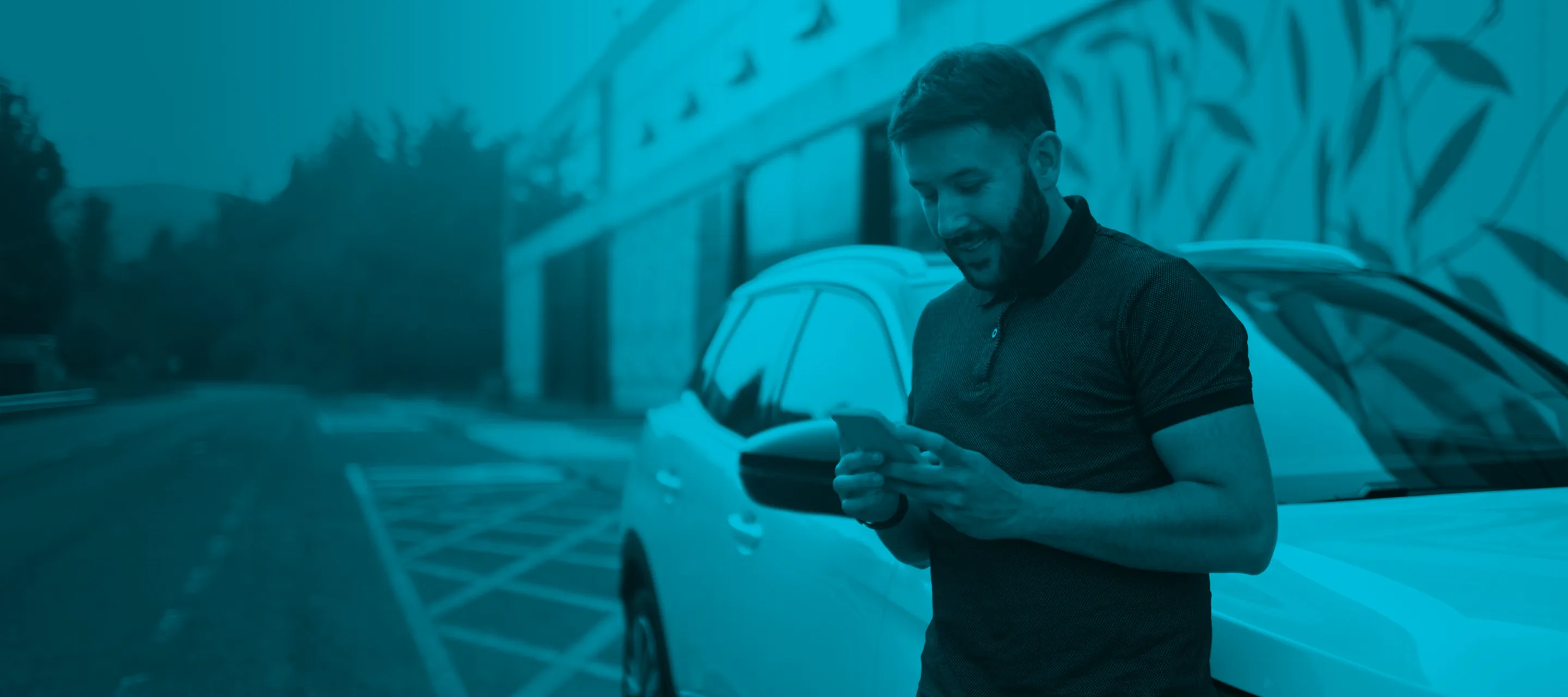 A man standing next to a car, looking at his smartphone and smiling.