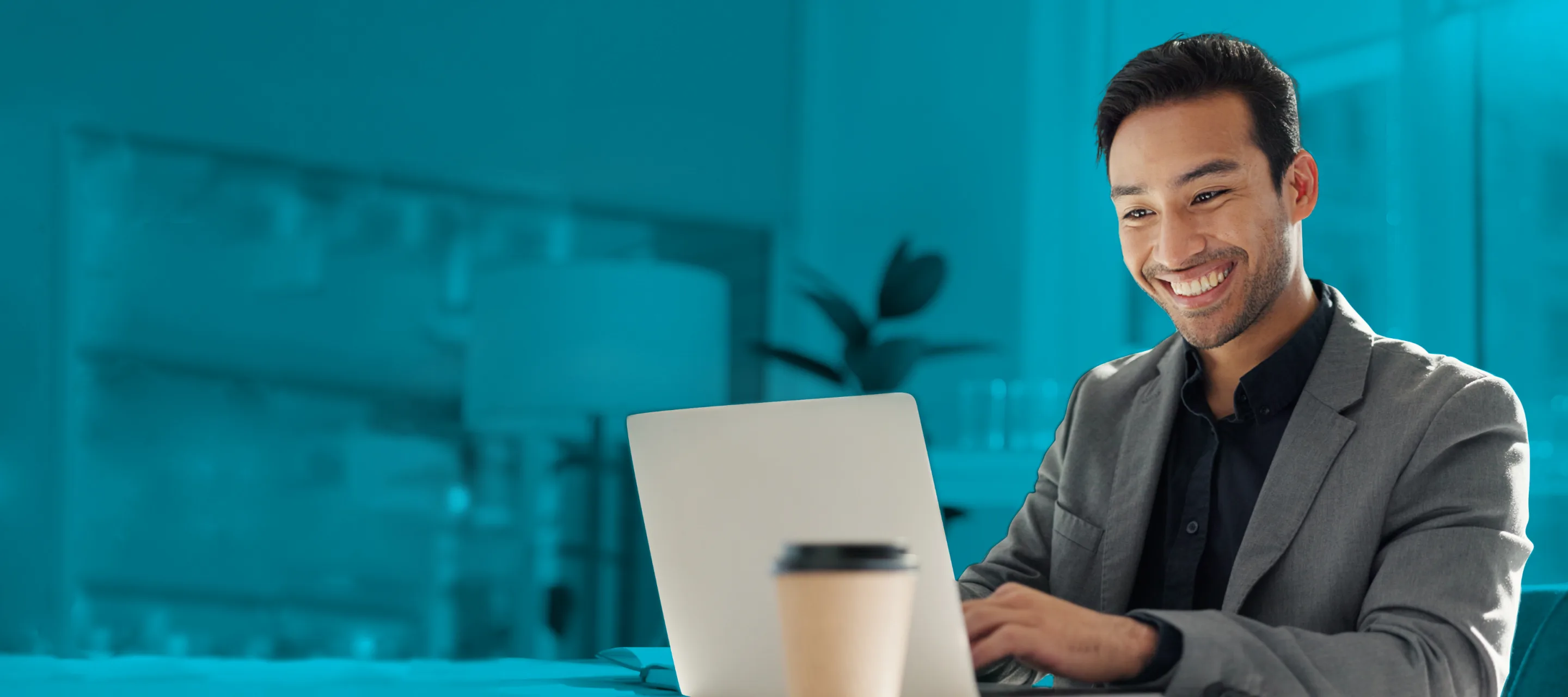 A man in a business suit smiles while working on a laptop, with a coffee cup placed beside him on the desk, set against the backdrop of a modern office environment.