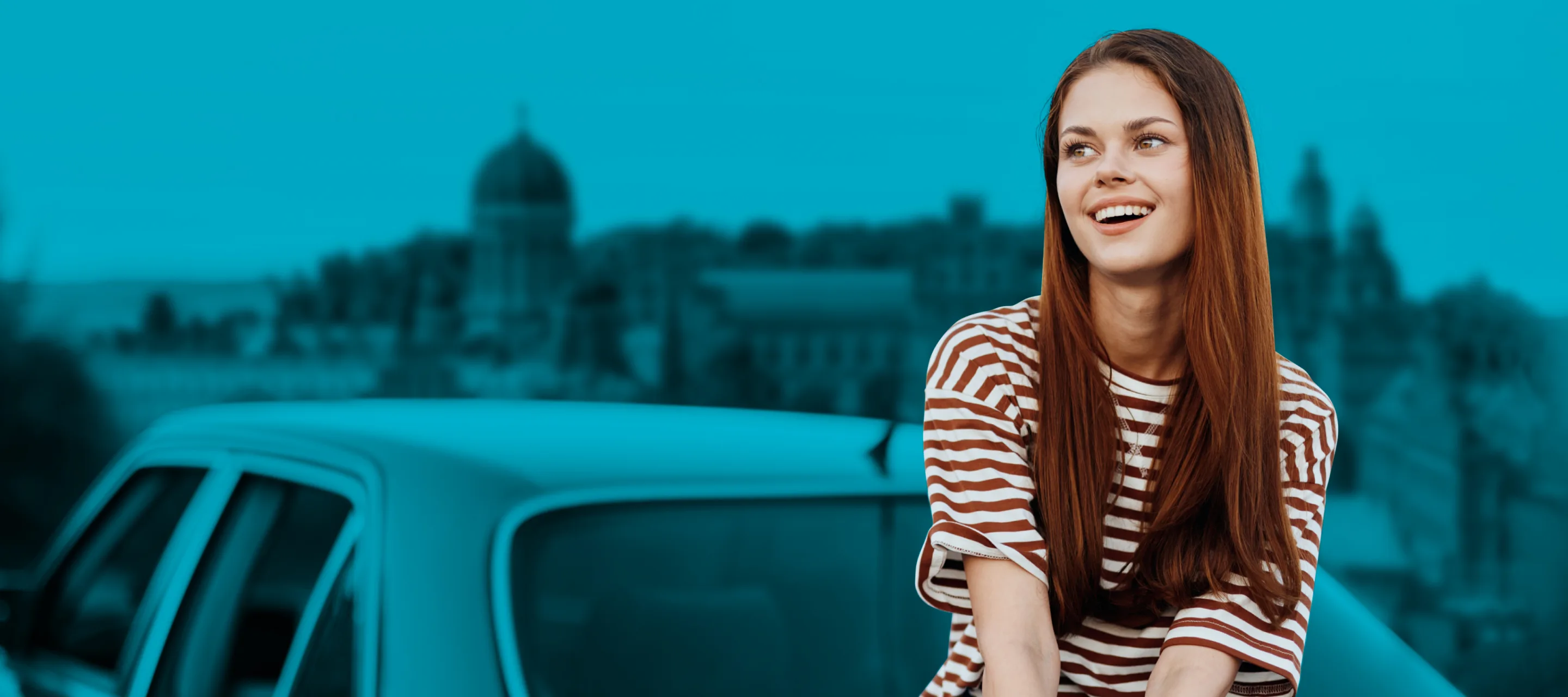 Smiling young woman with long brown hair sitting outdoors in a parking area, wearing a striped shirt.