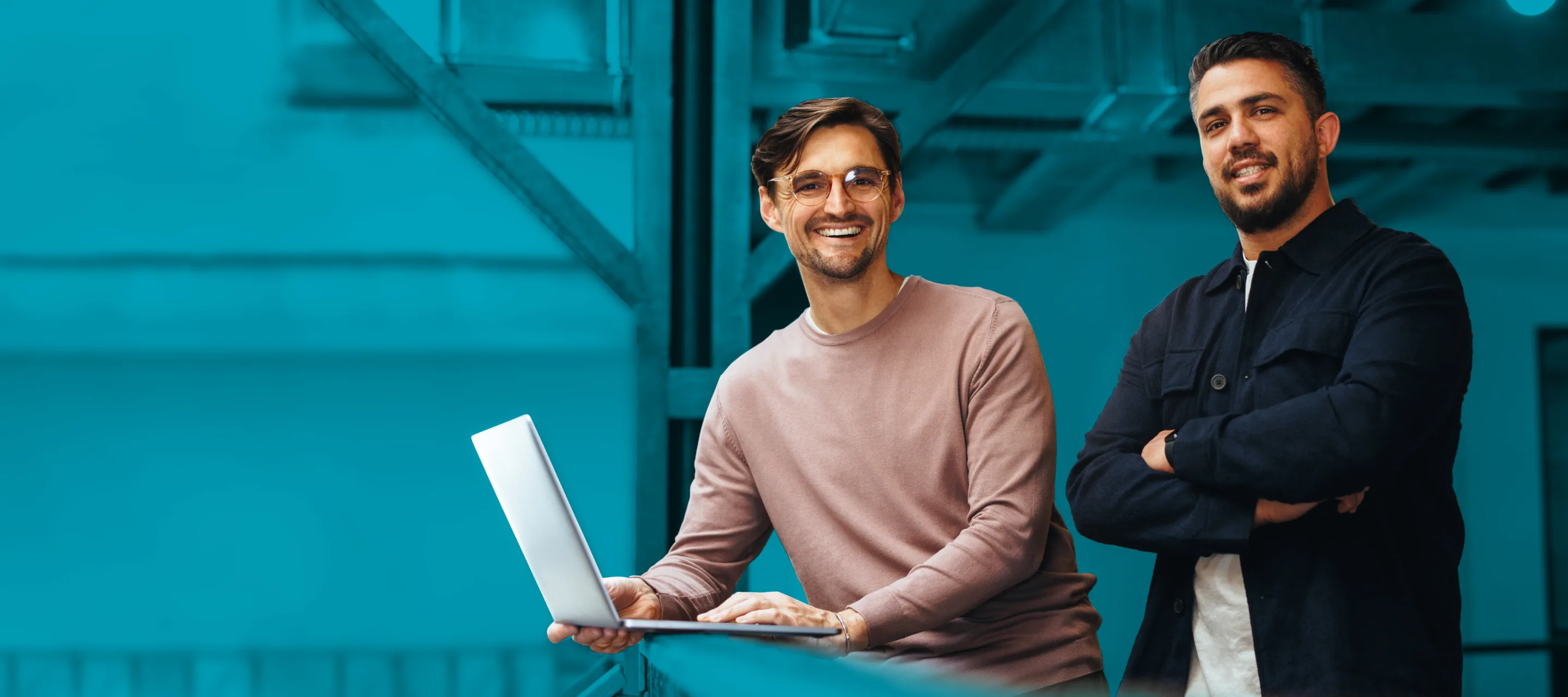 Two men are posing together in a modern office setting. One is holding a laptop and smiling, while the other stands beside him with his arms crossed, also smiling.