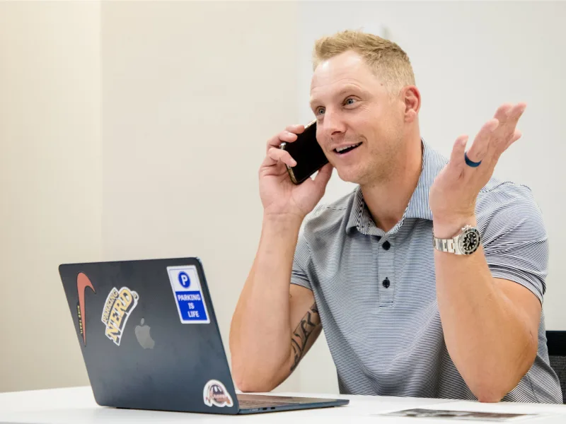 A man is sitting at a desk, talking on the phone while gesturing with his hand.