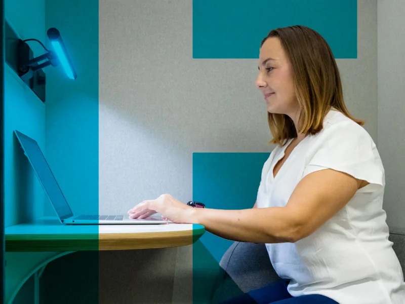 A woman is sitting at a small desk, working on a laptop in a well-lit, cozy workspace.