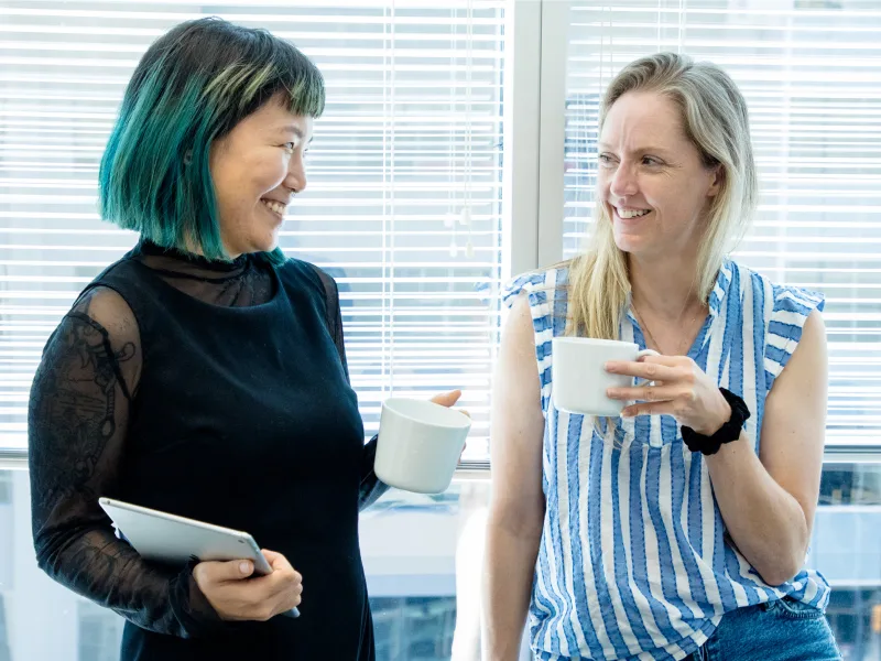 Two women are standing and smiling at each other, each holding a coffee cup.