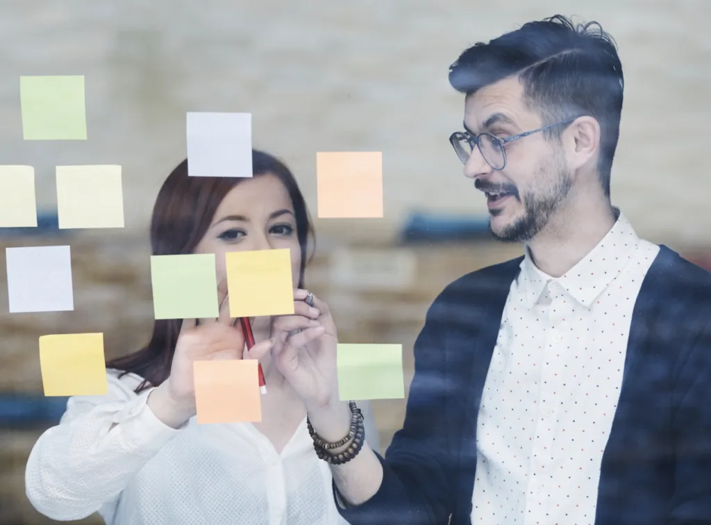 A man and woman are brainstorming together, placing sticky notes on a glass wall.