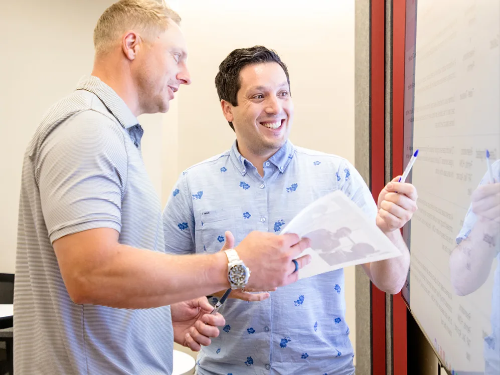 Two men are standing and discussing something on a whiteboard. One holds a pen and points to the board while the other holds a sheet of paper