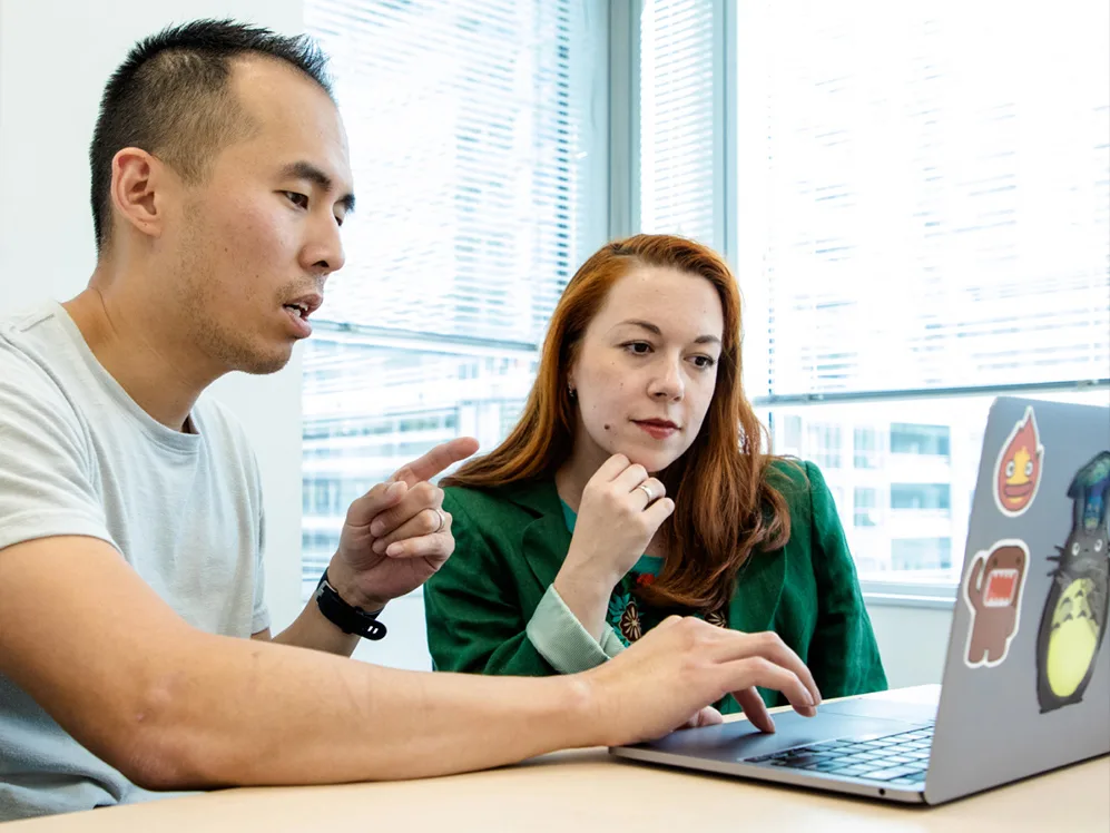 A man and woman are seated at a table, focused on a laptop.