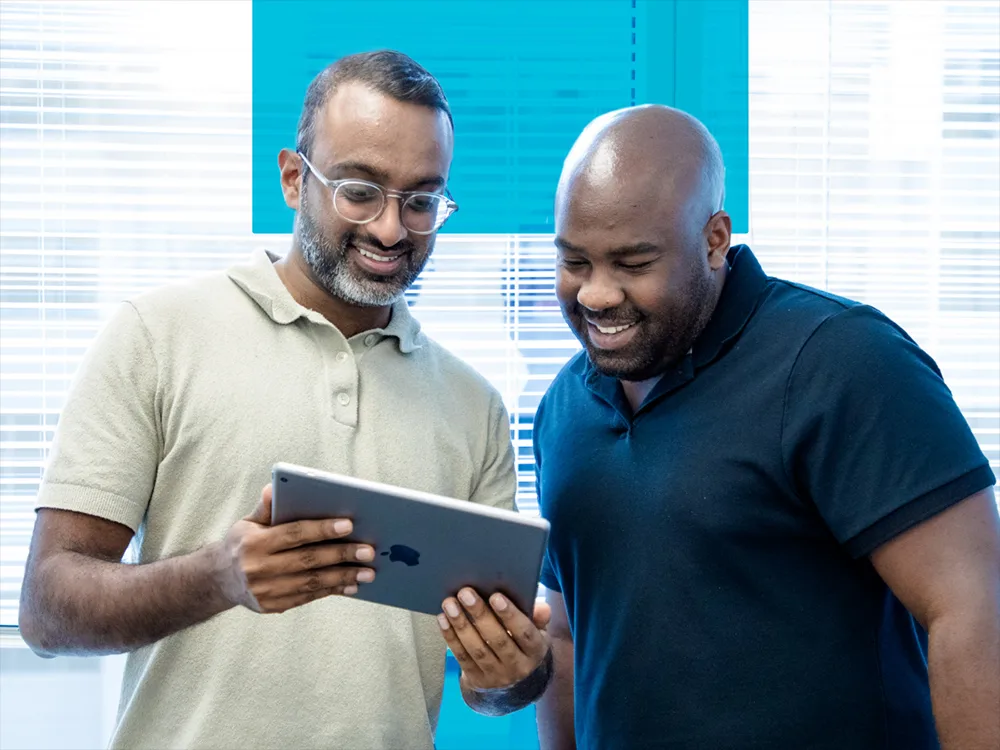 Two men smile while looking at a tablet together in an office setting.