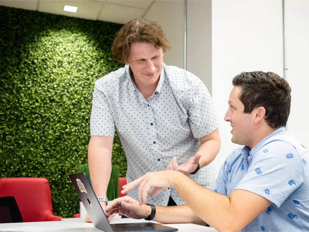 Two men in casual shirts are discussing something while looking at a laptop in an office setting