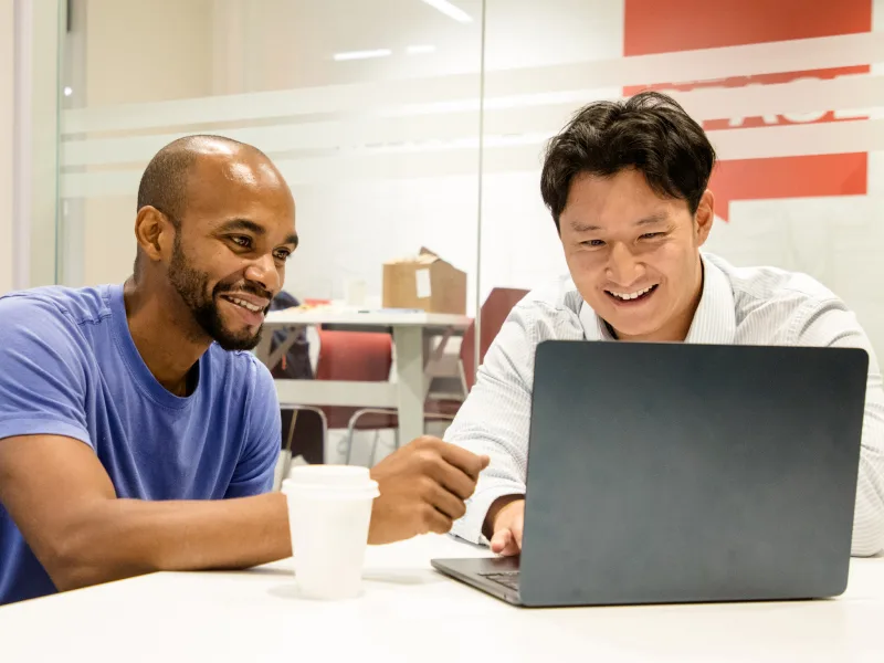 Two men smiling while looking at a laptop in an office setting.