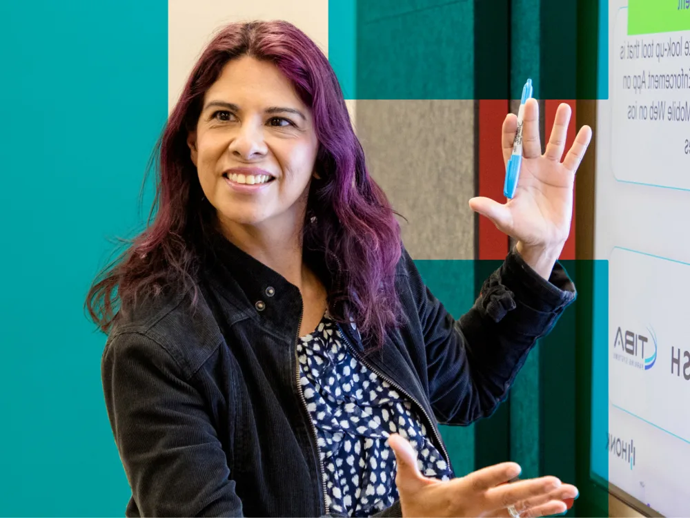 A woman with purple hair gestures while presenting in front of a screen.