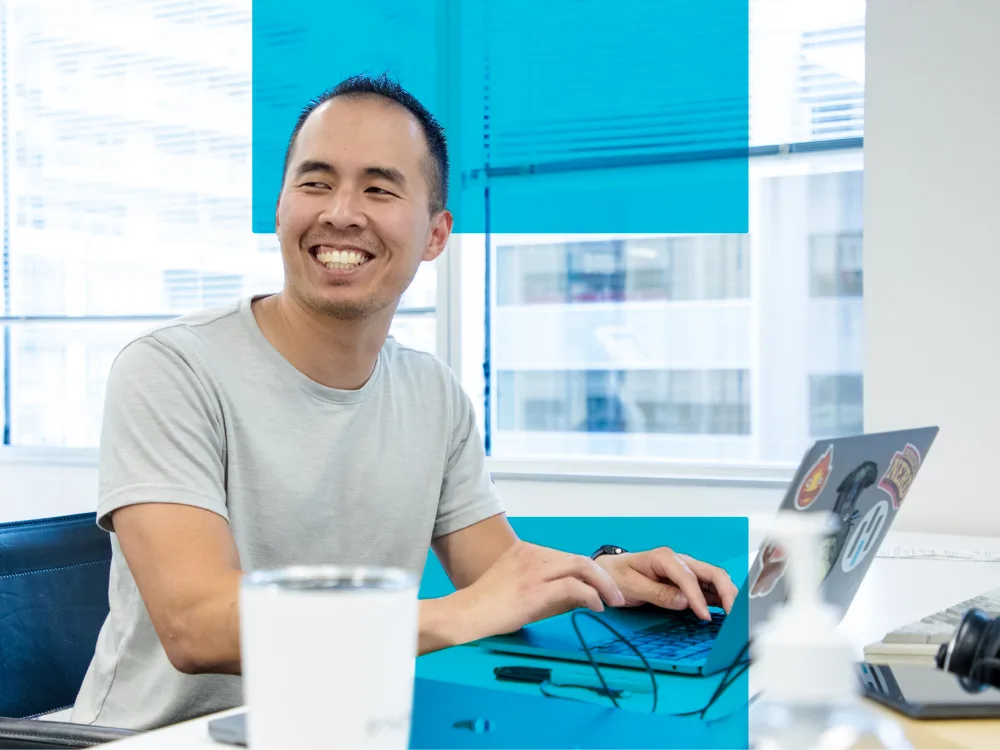 A man in a light gray t-shirt is smiling while typing on a laptop, seated at a desk in a bright office setting with large windows.