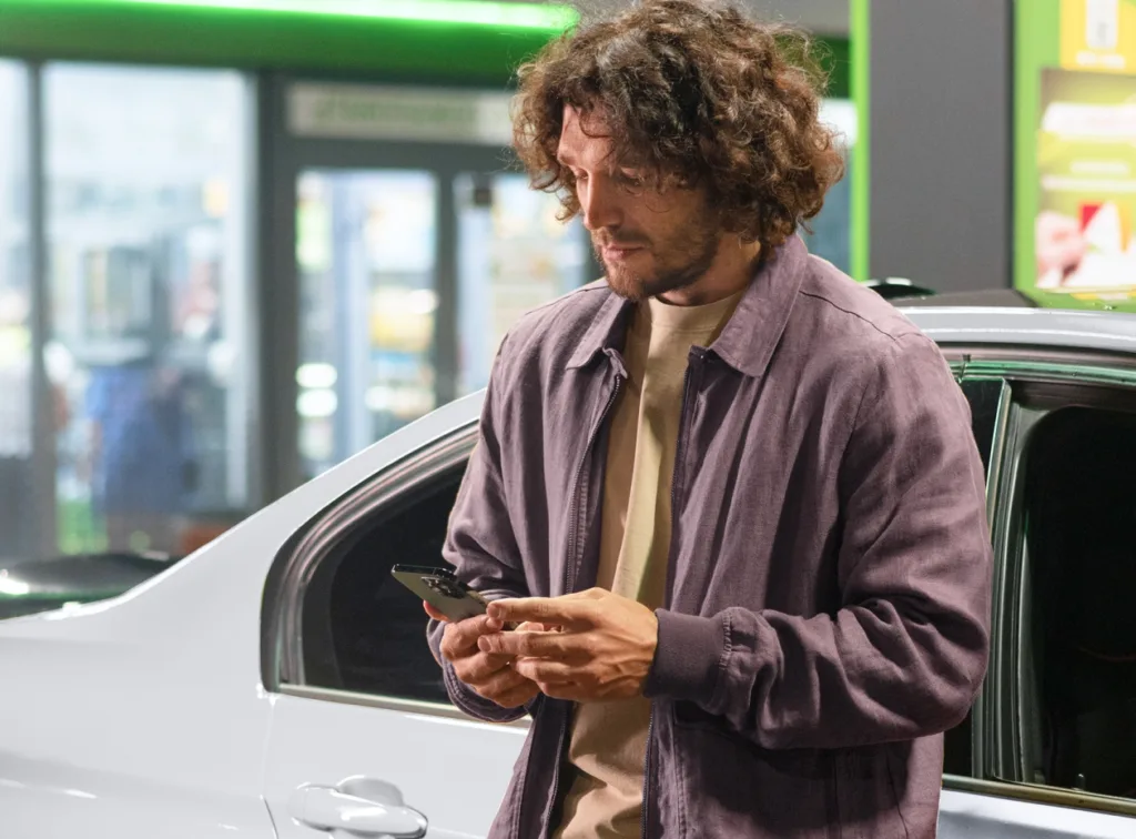 Man standing next to his car in a parking lot, using his phone.