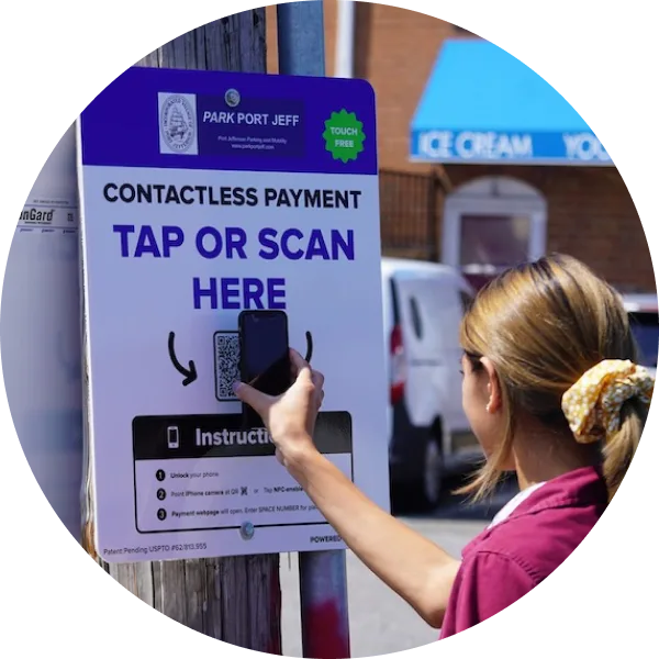 A woman holds her phone up to scan a QR code on a contactless payment sign that reads 'TAP OR SCAN HERE' for parking at Park Port Jeff