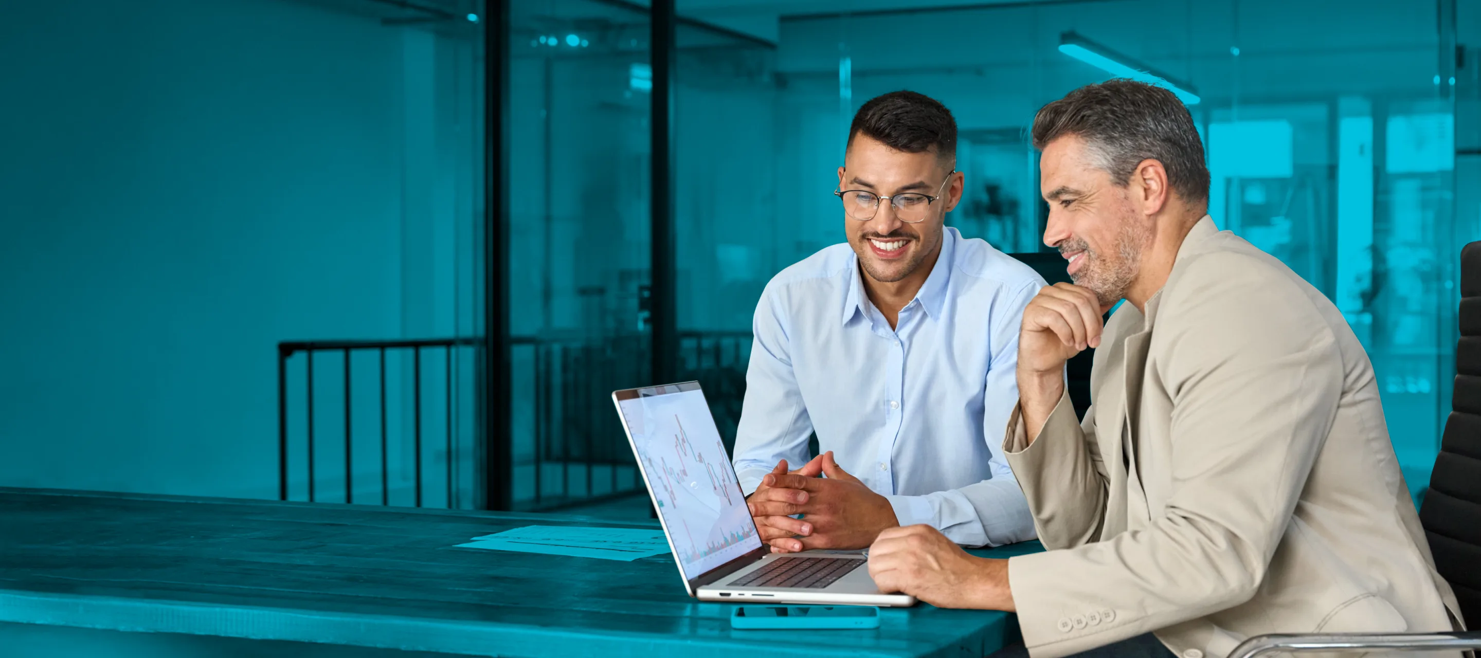 Two men sitting at a table in a modern office, smiling and engaged in a discussion while looking at a laptop.