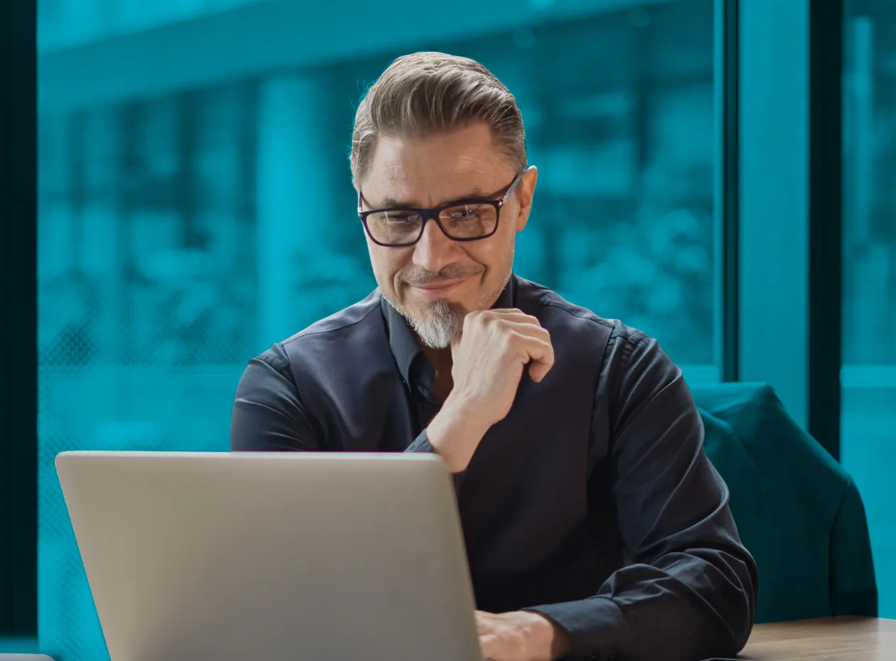 A man wearing glasses and a dark shirt sits at a desk, looking at a laptop with a thoughtful expression.