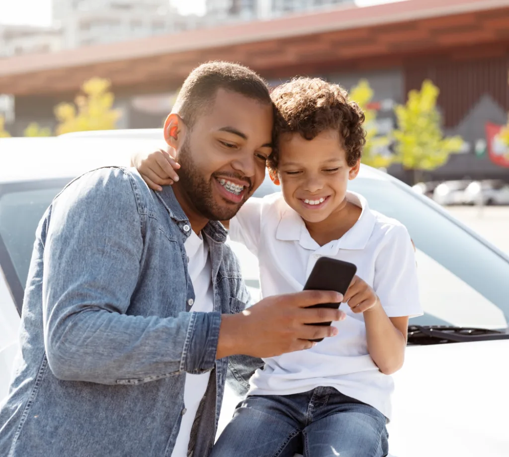 A smiling man and a young boy sit on the hood of a car outdoors, sharing a joyful moment as they look at a smartphone together.