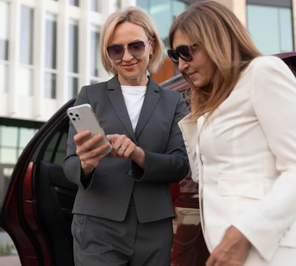 Two women dressed in business attire are standing next to a car, one holding a smartphone and showing something to the other.