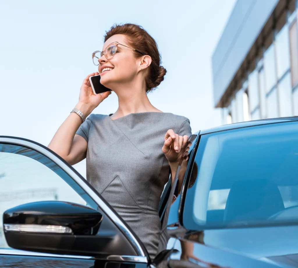 A woman dressed in business attire stands beside an open car door while talking on the phone.