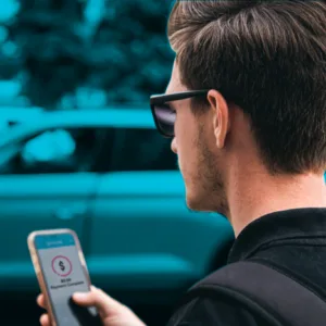 A man wearing sunglasses and a backpack is seen from behind, holding a smartphone and looking at an app related to parking.