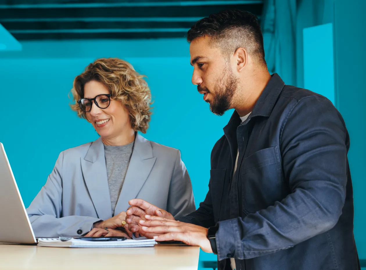 A man and woman, both in business attire, are standing at a desk, smiling and focused on a laptop, collaborating in a professional environment.