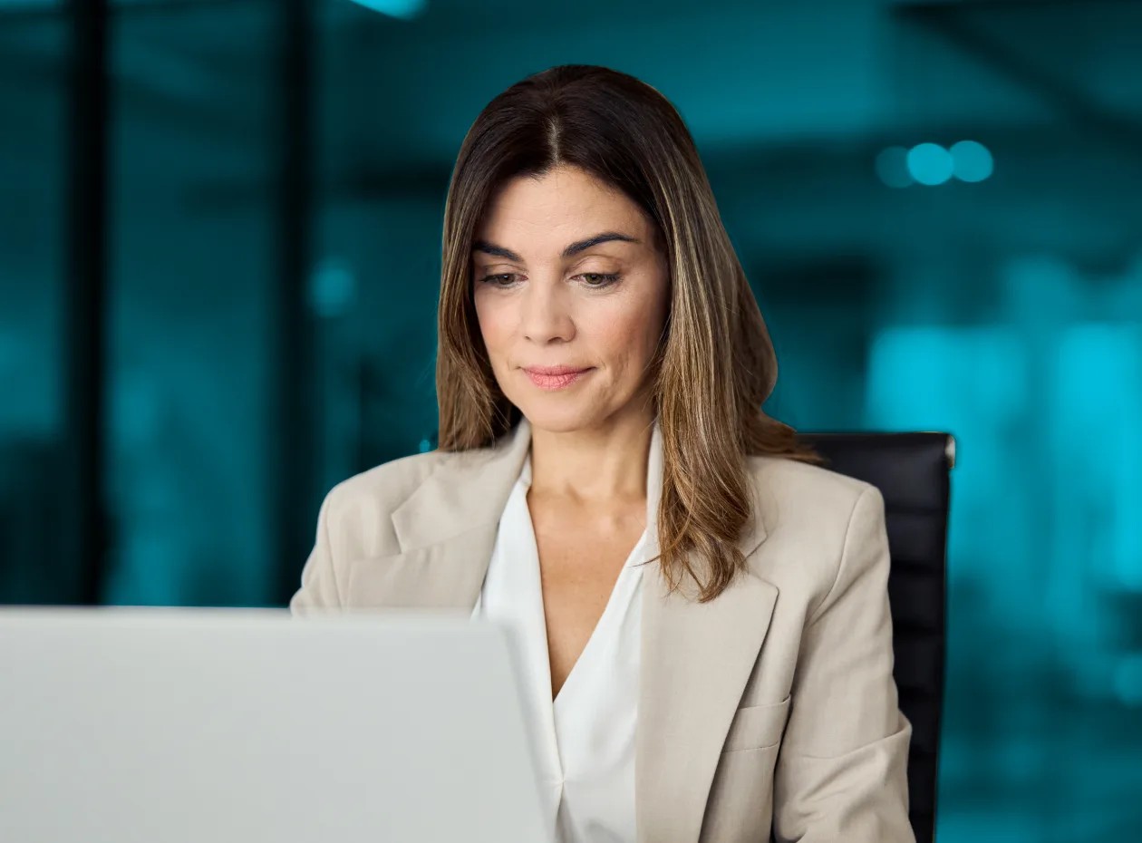 A woman in a light-colored business suit is seated at a desk, focused on her laptop screen.