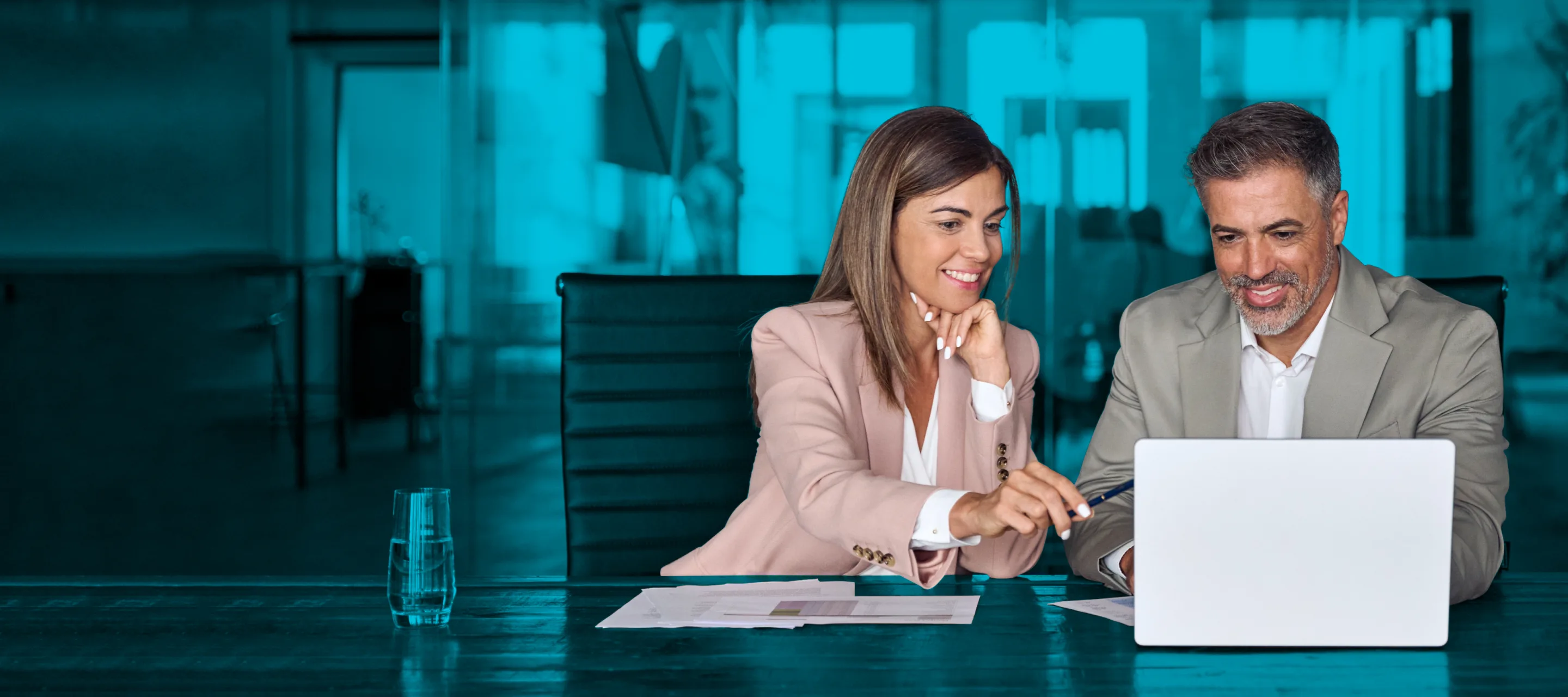 A man and woman smile while working together on a laptop in an office setting.